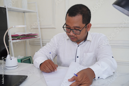 Focused young businessman signing up the documents of employment while working on laptop in office. Foreman manager CEO doing paperwork, working with digital data and statistics remotely photo