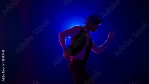 Canvas Print Man musician in stylish hat dancing on dark concert stage against blue spotlight and singing looking at camera, isolated on black studio background