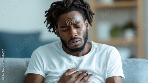 A distressed man wearing a white shirt holds his chest, appearing in discomfort, conveying a sense of urgency and discomfort amid a softly furnished background. photo