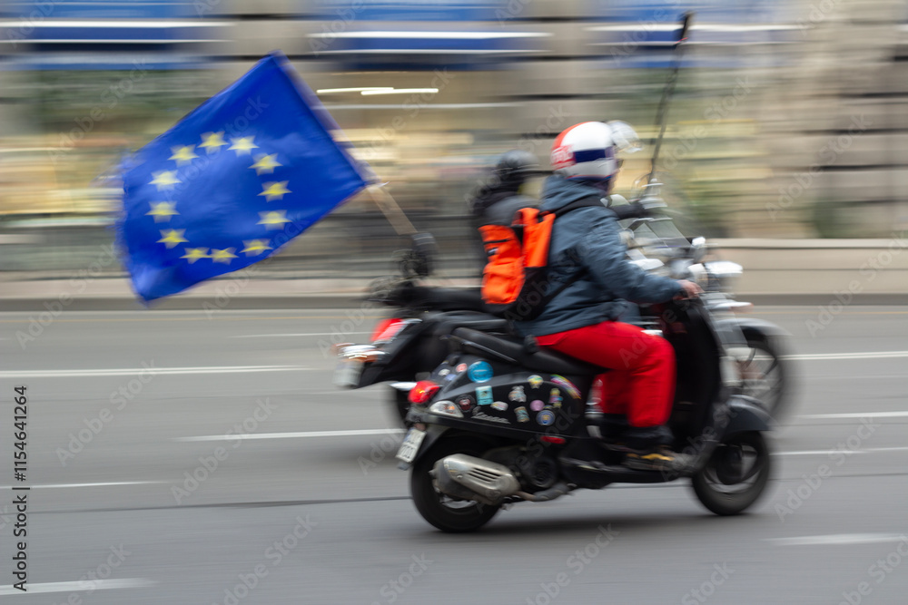 Fototapeta premium Unidentifiable man in red jacket in out of focus on motorcycle with EU flag with yellow stars. Street blurry defocused photo.Concept of transport, rally on city street, protest. Blurred background