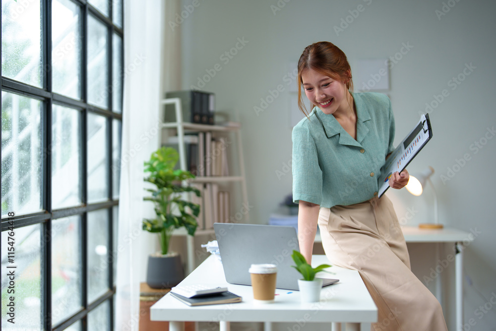 Asian businesswoman stands holding a file, checking financial reports, income, taxes, personal business at her desk. Checking data on laptop computer while standing at modern office. Asian businesswoman stands holding a file, checking financial reports, income, taxes, personal business at her desk. Checking data on laptop computer while standing at modern office.