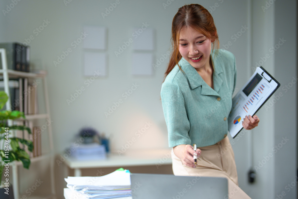 Asian businesswoman stands holding a file, checking financial reports, income, taxes, personal business at her desk. Checking data on laptop computer while standing at modern office. Asian businesswoman stands holding a file, checking financial reports, income, taxes, personal business at her desk. Checking data on laptop computer while standing at modern office.