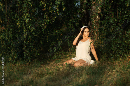 a girl sits on the grass in a forest or park. She is wearing a light dress and holds one hand to her head. There is a lot of greenery around her, including hanging tree branches with leaves.