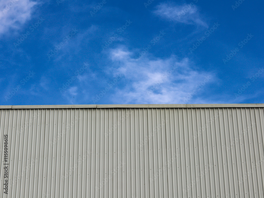 Industrial factory building with aluminum metal texture on the exterior wall against the background of blue sky and clouds, modern architecture of corrugated steel facade. Copyspace, design. Industrial factory building with aluminum metal texture on the exterior wall against the background of blue sky and clouds, modern architecture of corrugated steel facade. Copyspace, design.