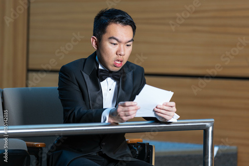 A man in a tuxedo sits in an auditorium holding script photo