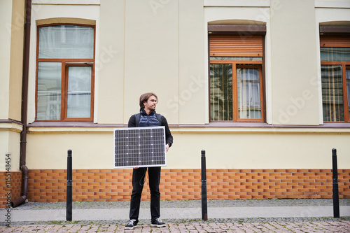 Man holding solar panel, stands in front of old building. Guy in a black hoodie and navy blue quilted vest. Aged architecture contrasts with modernity of renewable energy. photo