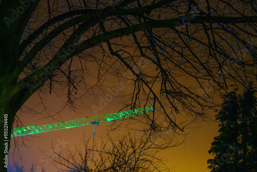green lit construction crane with orange sky in background and bare branches in the foreground in early morning