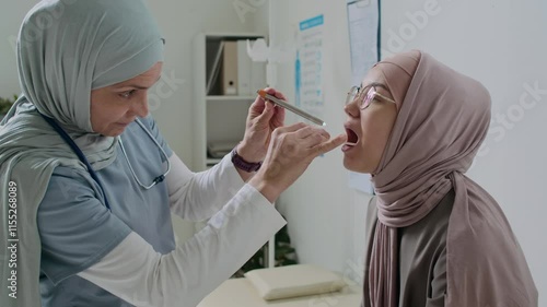 Female doctor in hijab performing oral examination with tongue depressor and penlight on Muslim patient in clinic photo