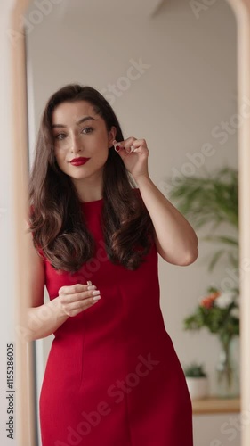 Young elegant woman in a red dress choosing between two earrings, looking at her reflection in a full-length mirror.
