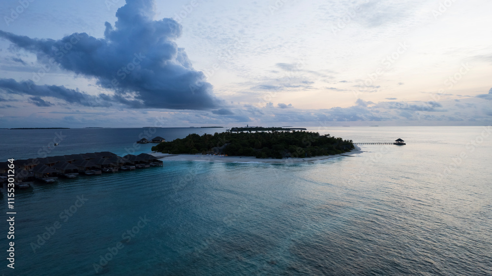 Top view of the sunset over an island in the ocean. Large dark clouds with a orange tinge. Turquoise ocean water, white sand of the beach. Green palm trees. Villas on the water. Water is like a mirror