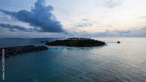 Obraz na płótnie Top view of the sunset over an island in the ocean. Large dark clouds with a orange tinge. Turquoise ocean water, white sand of the beach. Green palm trees. Villas on the water. Water is like a mirror w salonie
