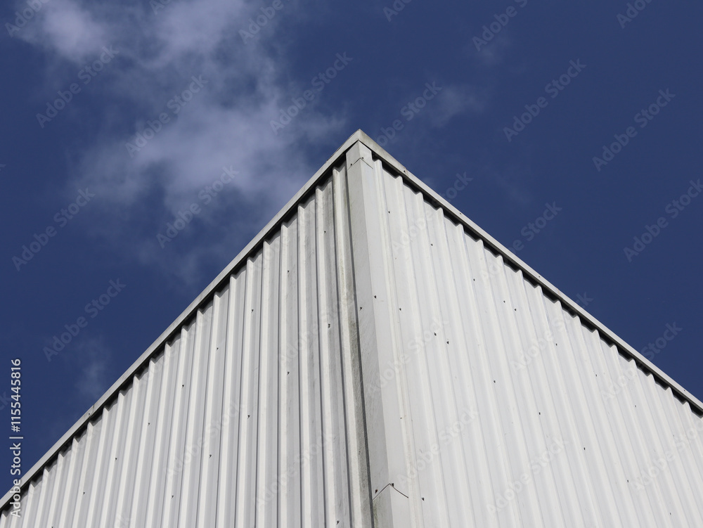 Industrial factory building with aluminum metal texture on the exterior wall against the background of blue sky and clouds, modern architecture of corrugated steel facade. Copyspace, design. Industrial factory building with aluminum metal texture on the exterior wall against the background of blue sky and clouds, modern architecture of corrugated steel facade. Copyspace, design.