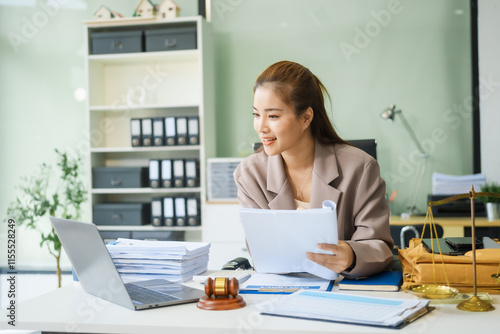 A young female lawyer sits at her desk in the office, providing online legal advice professionally. Legal documents, a computer, a judge's gavel, and scales symbolize justice and law