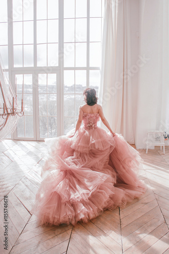 young girl with short black hair in pink princess dress with train is standing from the back and looking cute near huge panoramic white window background at sunny day. fashion concept, free space