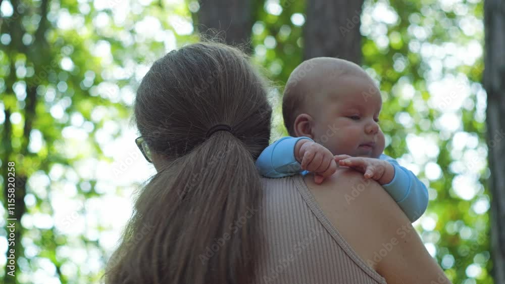 A woman in glasses gently cradles a small child to her breast, with a forest in background. The mother is standing with her back to camera, baby is looking at camera from behind her shoulder A woman in glasses gently cradles a small child to her breast, with a forest in background. The mother is standing with her back to camera, baby is looking at camera from behind her shoulder
