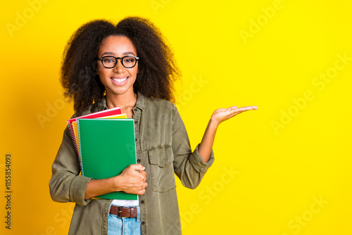 Photo of positive girl wear khaki shirt in glasses holding copybooks arm presenting object empty space isolated on yellow color background photo