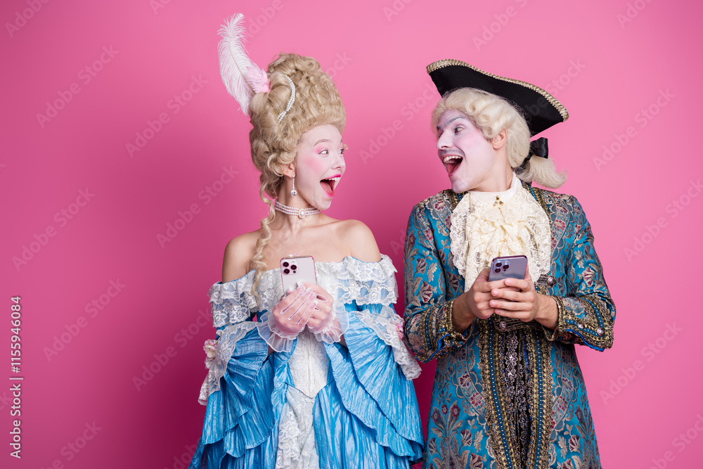 Joyful couple in elegant baroque costumes sharing a moment on their phones against a vibrant pink backdrop Joyful couple in elegant baroque costumes sharing a moment on their phones against a vibrant pink backdrop