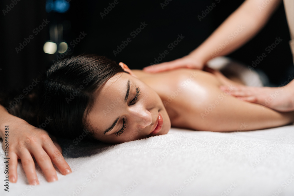 Close-up portrait of beautiful calm young woman client relaxing during back massage in spa salon with dark interior. Professional massage therapist providing therapeutic back massage to relaxed female Close-up portrait of beautiful calm young woman client relaxing during back massage in spa salon with dark interior. Professional massage therapist providing therapeutic back massage to relaxed female
