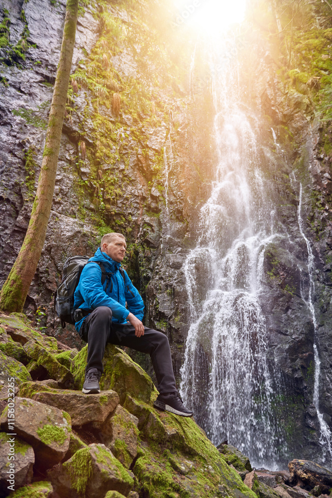 Tourist attraction of Germany - falls of Burgbach Waterfall near Schapbach, Black Forest, Baden-Wurttemberg, Germany. Man hiker in blue jacket standing on stone and looks at flow of falling water Tourist attraction of Germany - falls of Burgbach Waterfall near Schapbach, Black Forest, Baden-Wurttemberg, Germany. Man hiker in blue jacket standing on stone and looks at flow of falling water