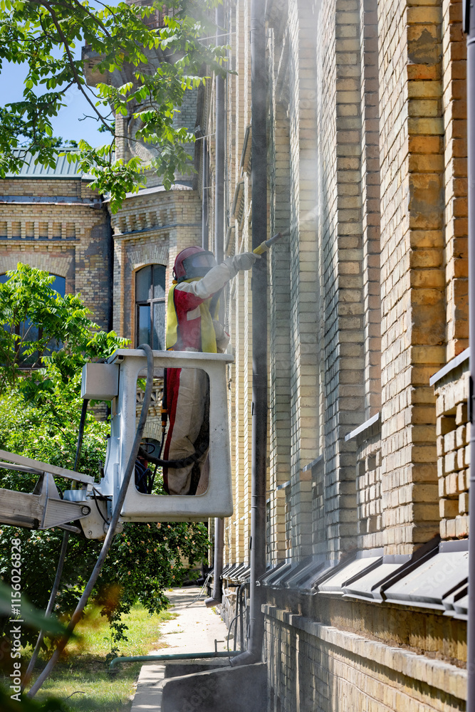Worker on an elevating platform cleaning the facade of an old brick building during sunny weather