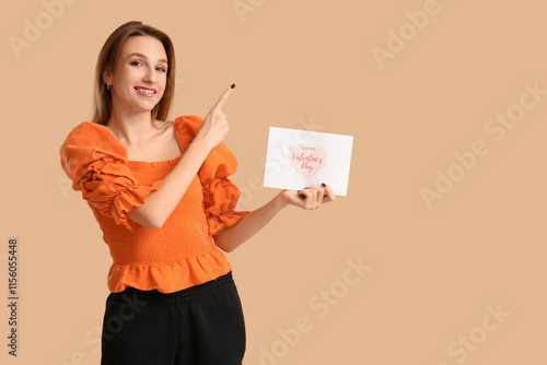 Beautiful young woman holding greeting card with text HAPPY VALENTINE'S DAY and pointing at something on beige background