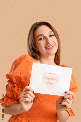 Happy young woman holding greeting card with text HAPPY VALENTINE'S DAY on beige background