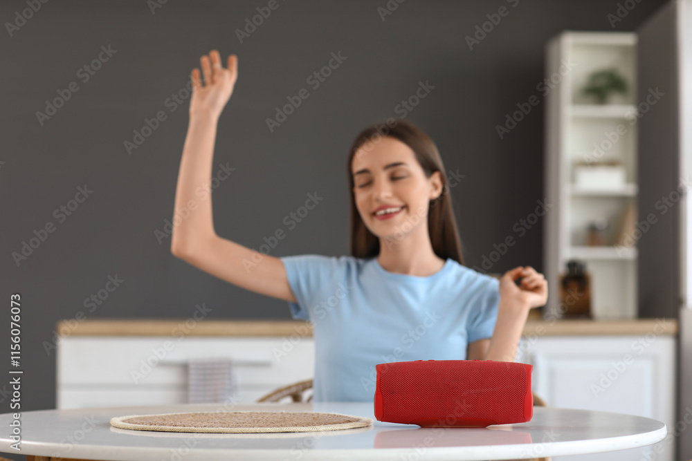 Beautiful young happy woman with wireless portable speaker sitting at table in kitchen Beautiful young happy woman with wireless portable speaker sitting at table in kitchen