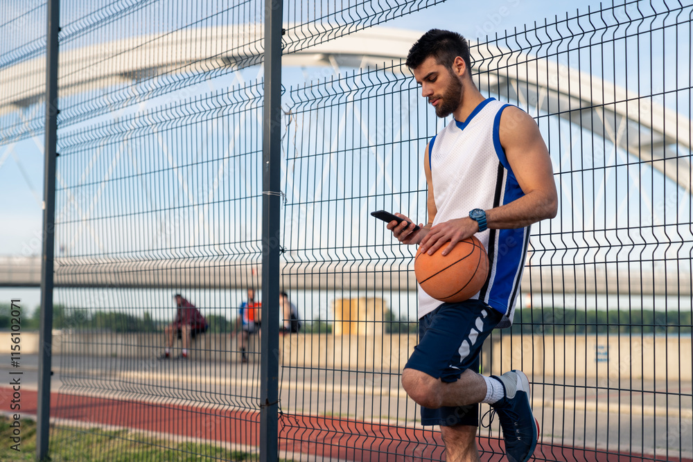 Young Caucasian male athlete in basketball attire checks smartphone while holding basketball near mesh fence on outdoor sports court. Young Caucasian male athlete in basketball attire checks smartphone while holding basketball near mesh fence on outdoor sports court.