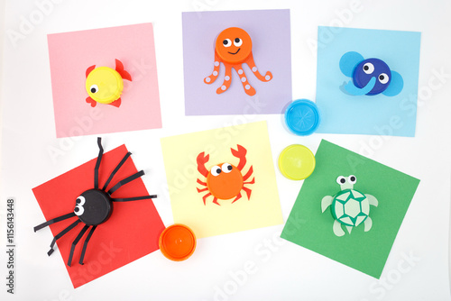 Animal crafts using bottle caps and colored paper on a white background. Flat lay composition with fish, octopus, elephant, spider, crab, and turtle designs. DIY kids activity and recycling concept, photo