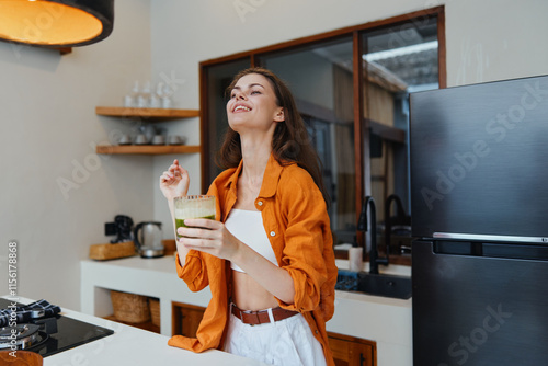 Young woman in orange shirt enjoying a healthy green drink in a modern kitchen, radiating happiness and wellness, vibrant colors, bright interior design