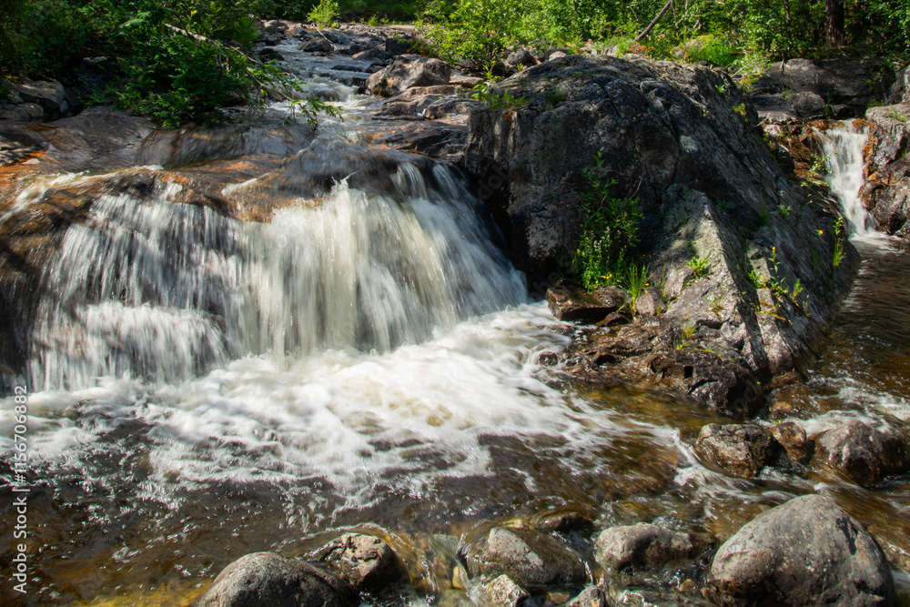 Naklejka premium Norwegian mountain river with fast flowing clear and sparkling water and a waterfall that forms white foam on rocks surrounded by green coniferous trees. Sunny summer day.