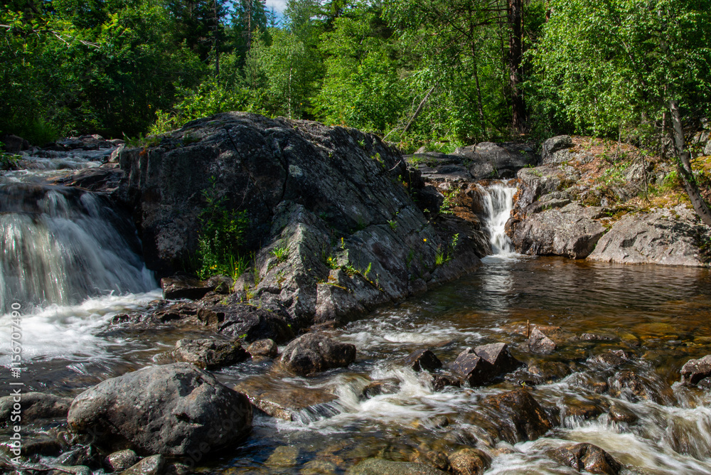 Fototapeta premium Norwegian mountain river with fast flowing clear and sparkling water and a waterfall that forms white foam on rocks surrounded by green coniferous trees. Sunny summer day.