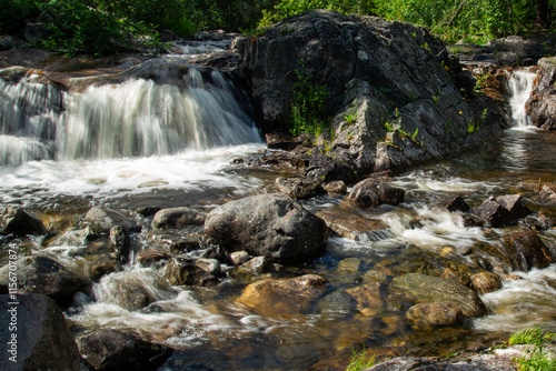 Norwegian mountain river with fast flowing clear and sparkling water and a waterfall that forms white foam on rocks surrounded by green coniferous trees. Sunny summer day.