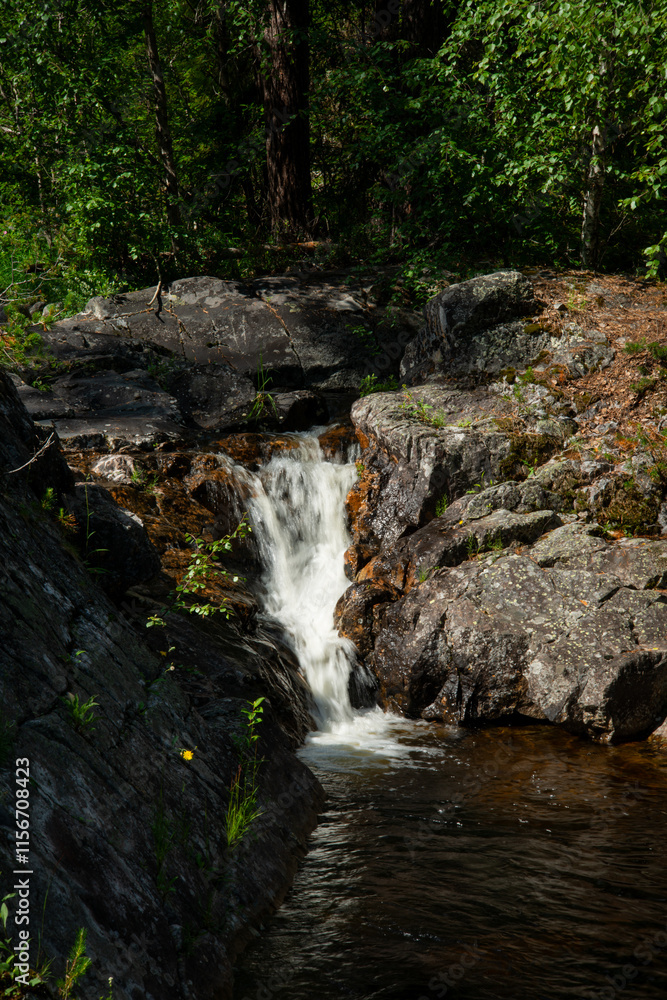 Norwegian mountain river with fast flowing clear and sparkling water and a waterfall that forms white foam on rocks surrounded by green coniferous trees. Sunny summer day. Norwegian mountain river with fast flowing clear and sparkling water and a waterfall that forms white foam on rocks surrounded by green coniferous trees. Sunny summer day.