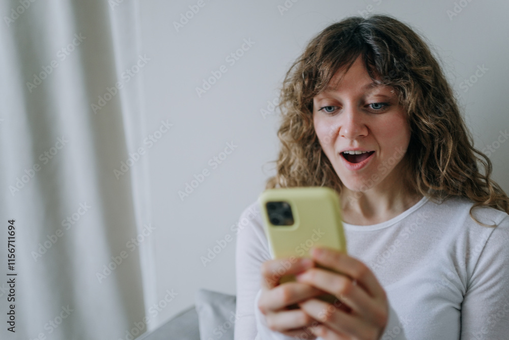 Young woman with curly hair holding a yellow smartphone and looking surprised by the news she is reading while sitting on a sofa at home Young woman with curly hair holding a yellow smartphone and looking surprised by the news she is reading while sitting on a sofa at home