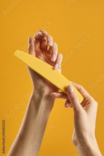 Hands gently filing nails with a yellow nail file against a bright orange background during a self-care session at home