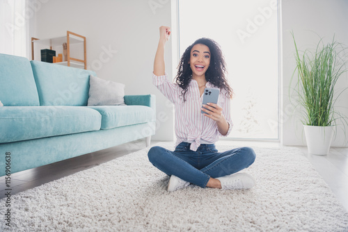 Full size photo of pretty young girl sit floor hold phone raise fist weekend white interior apartment indoors photo