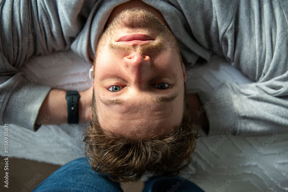 Fototapeta premium Chill male with blue eyes looking into camera candid grey jumper in bed relaxed, has an ear bud in an ear with hands behind his head. Comfortable on white textured bedding smiling and happy.