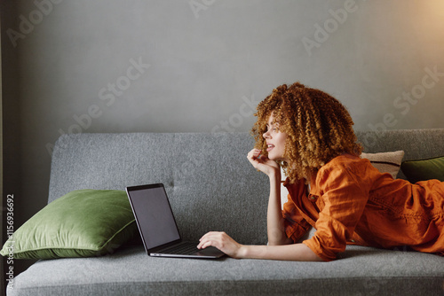 Fotografie Young woman with curly hair in orange blouse working on laptop at home with gree