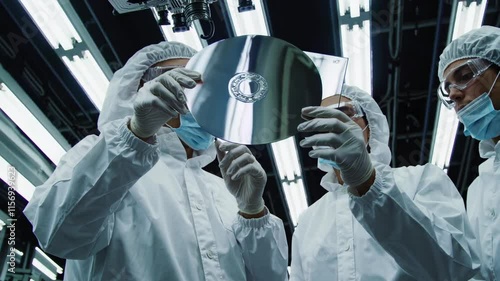 Personnel analyze a semiconductor wafer under bright laboratory lights in a cleanroom environment