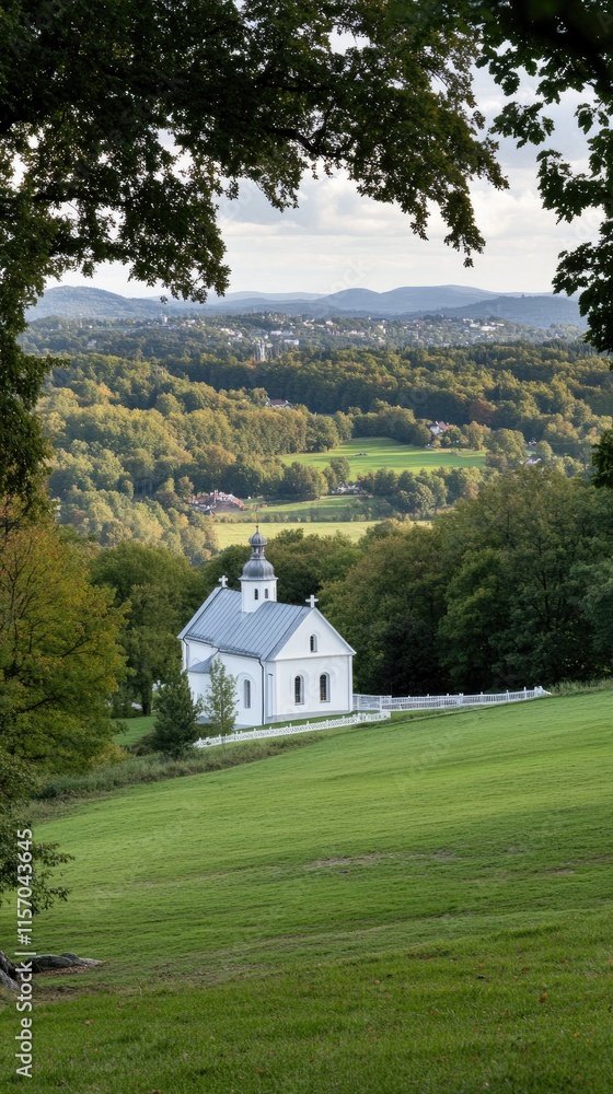 Obraz premium A quaint white and gray church with a black roof sits in a green field, surrounded by trees displaying autumn colors under a cloudy sky