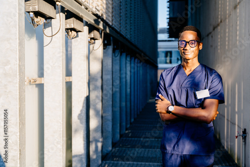 African American male doctor in blue medical uniform outside clinic. Smiling young man medical specialty posing for photo outdoors. Cheerful person with arms crossed. photo