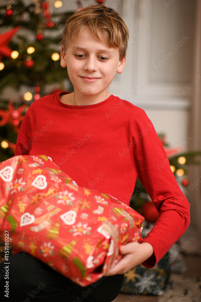 Smiling boy holding christmas present sitting near decorated christmas tree Smiling boy holding christmas present sitting near decorated christmas tree
