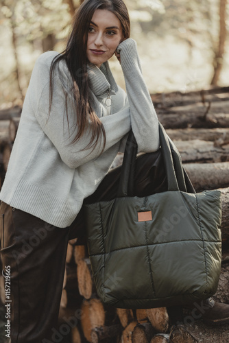 A thoughtful woman in a sweater with a large, quilted bag poses against the background of a pile of wood in natural, subdued lighting. (selective focus)