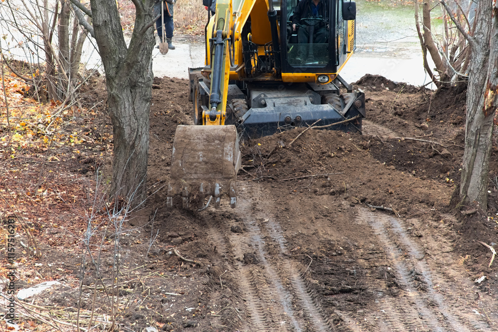 The excavator bucket reaches down to begin the first stage of soil extraction on a site with trees, excavator operation in construction during autumn. The excavator bucket reaches down to begin the first stage of soil extraction on a site with trees, excavator operation in construction during autumn.
