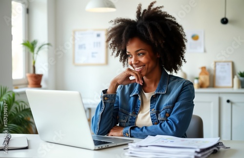 Happy young African American woman working on laptop in cozy studio. Looking at computer screen with smile. Appears focused, engaged in task related to real estate investment data analysis, financial photo