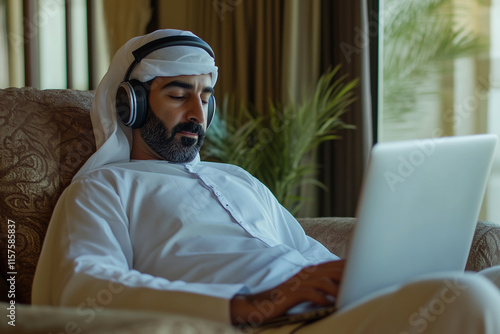  man wearing a white robe and a turban is sitting on a couch with a laptop in front of him. He is wearing headphones and he is listening to music photo