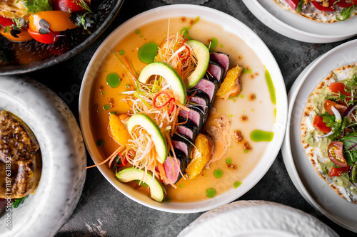 Fotografie beautifully presented dish featuring seared tuna slices garnished with fresh vegetables, citrus segments, and vibrant sauces, surrounded by an array of colorful appetizers on a stylish table setting