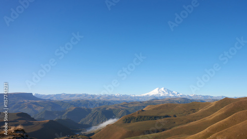 Panoramic picturesque view of the mountain landscape with morning fog in the valley. Snow-covered Elbrus in the distance against the blue sky. Copy space. photo