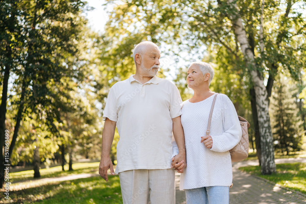 Low-angle view of charming elderly couple strolling in lush park, talking and smiling, embodying love and happiness in retirement, enjoying sunny day. Active senior male and female enjoying retirement Low-angle view of charming elderly couple strolling in lush park, talking and smiling, embodying love and happiness in retirement, enjoying sunny day. Active senior male and female enjoying retirement